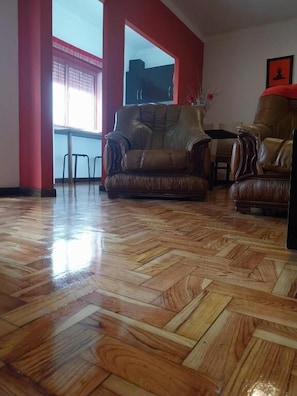 Una habitación con piso de madera pulida, dos sofás de cuero, una mesa de comedor y un ventanal con marco rojo.