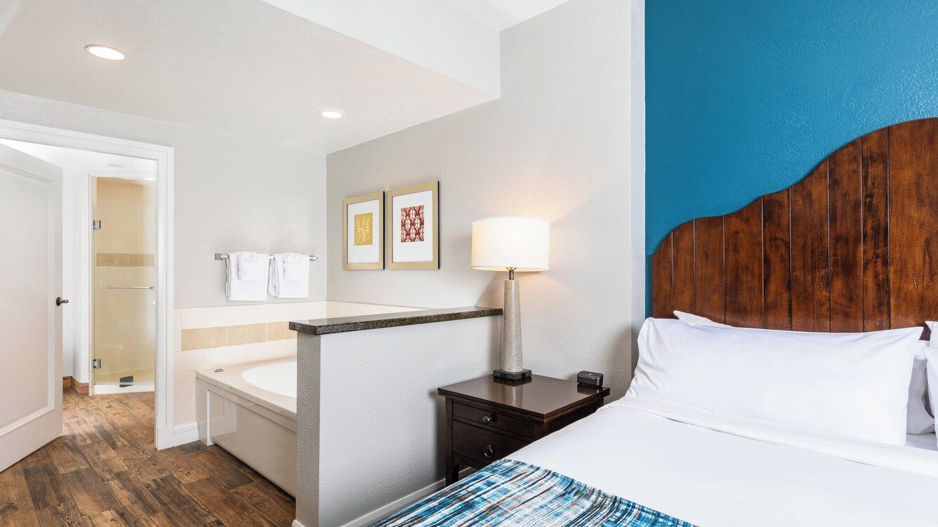 Quarto de hotel com uma cama, mesa de cabeceira, luminária, banheiro com chuveiro e porta que dá para outro cômodo.