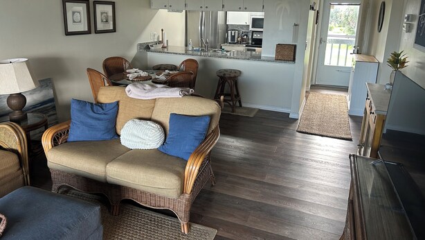 Ein Wohnzimmer mit einer Couch, Sesseln, einem Couchtisch, einer Küchenzeile mit Schränken und Geräten sowie Blick ins Freie.
