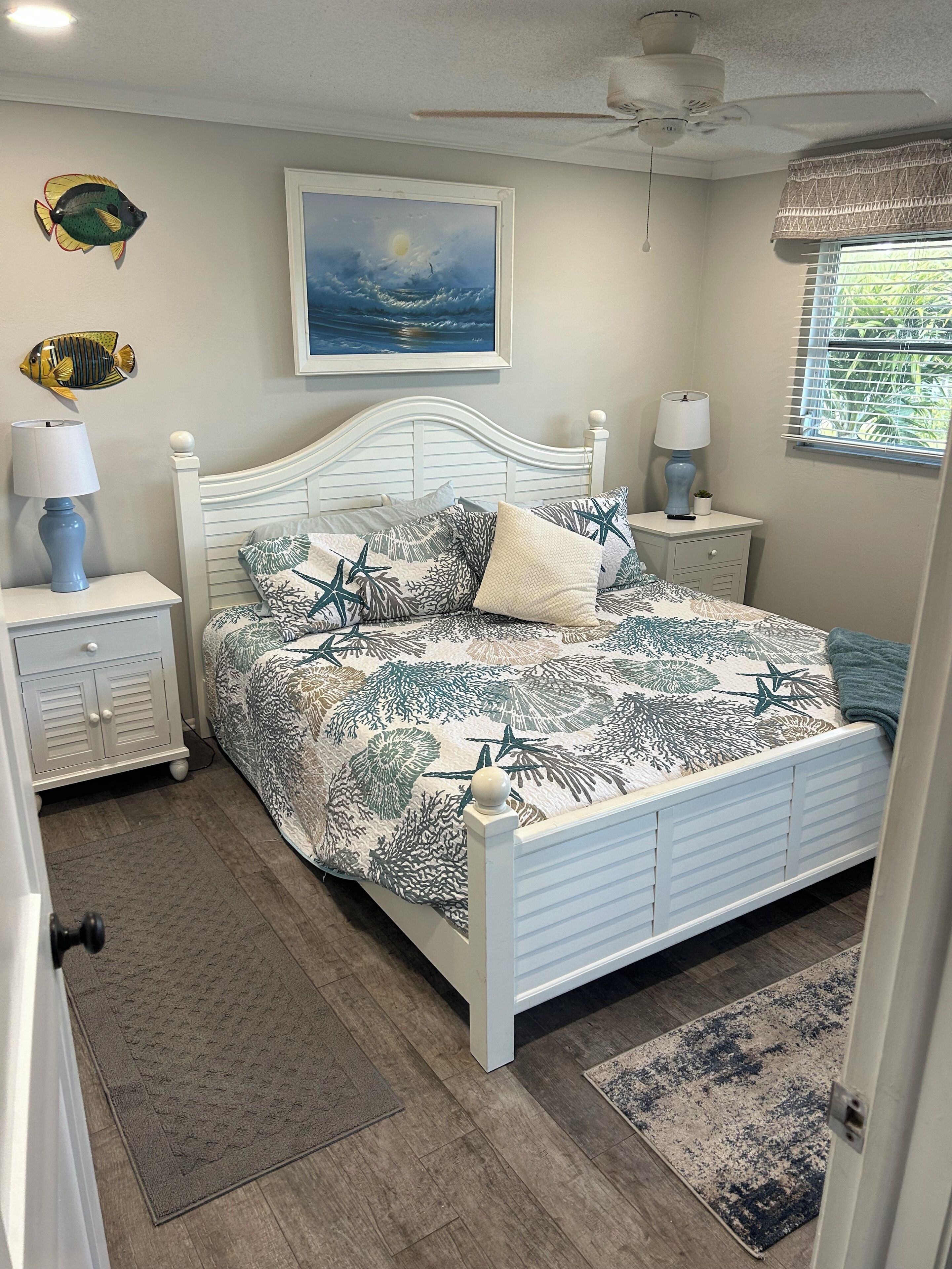 A bedroom with a white bed, a nightstand with a lamp, a wall-mounted fish decoration, a framed painting, and a window with blinds.