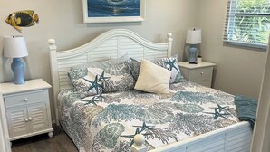 Ein Schlafzimmer mit einem weißen Bett, einem Nachttisch mit Lampe, einer Wanddekoration mit Fischmotiv, einem gerahmten Gemälde und einem Fenster mit Jalousien.