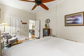 Ein Schlafzimmer mit einem großen Bett, einem Deckenventilator, Holzboden und Blick auf einen Raum mit einem Sessel und einer Lampe.
