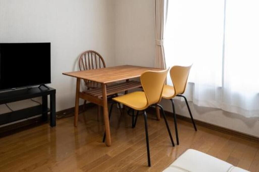 A room with a wooden dining table, two chairs, a television on a stand, and a window with curtains.