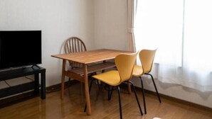 Une pièce comprenant une table à manger en bois, deux chaises, un téléviseur posé sur un meuble et une fenêtre avec des rideaux.