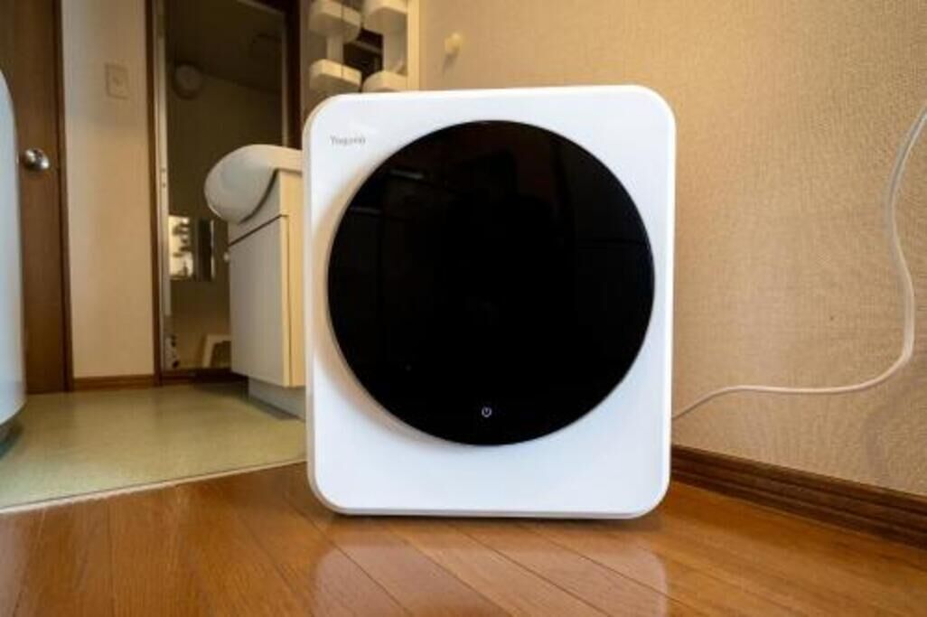 Un purificateur d’air portatif compact et moderne, de couleur blanche, avec une façade circulaire, posé sur un plancher en bois dans une pièce dont le mur est de couleur beige et où l’on aperçoit une porte.