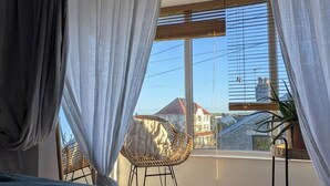 Une chambre avec une grande fenêtre, un fauteuil en osier et une vue sur les bâtiments environnants.
