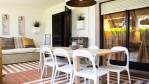Un comedor con mesa de madera y sillas blancas, sofá con cojines, suelo de ladrillo y un amplio ventanal con cortinas.