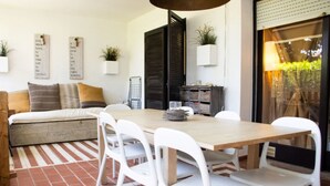 Un comedor con mesa de madera y sillas blancas, un sofá con cojines, un cerrojo negro y una ventana con vistas.