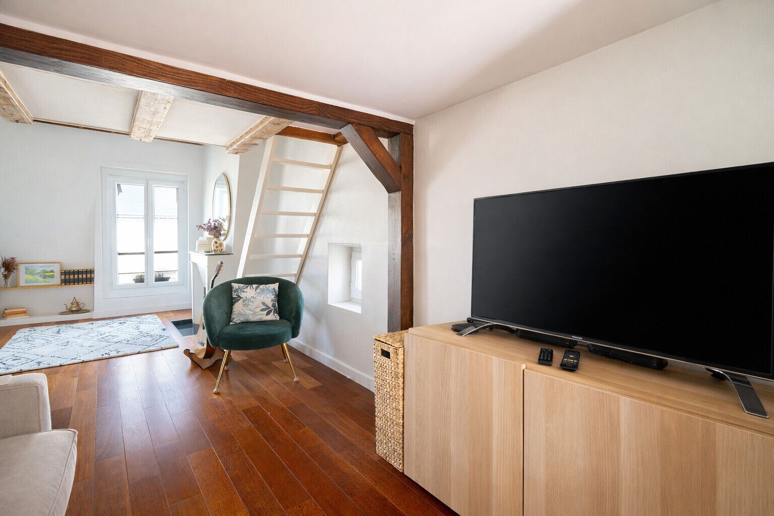 A living room with a large flat-screen TV on a wooden cabinet, a green armchair, a wooden floor, and a staircase leading to an upper level.