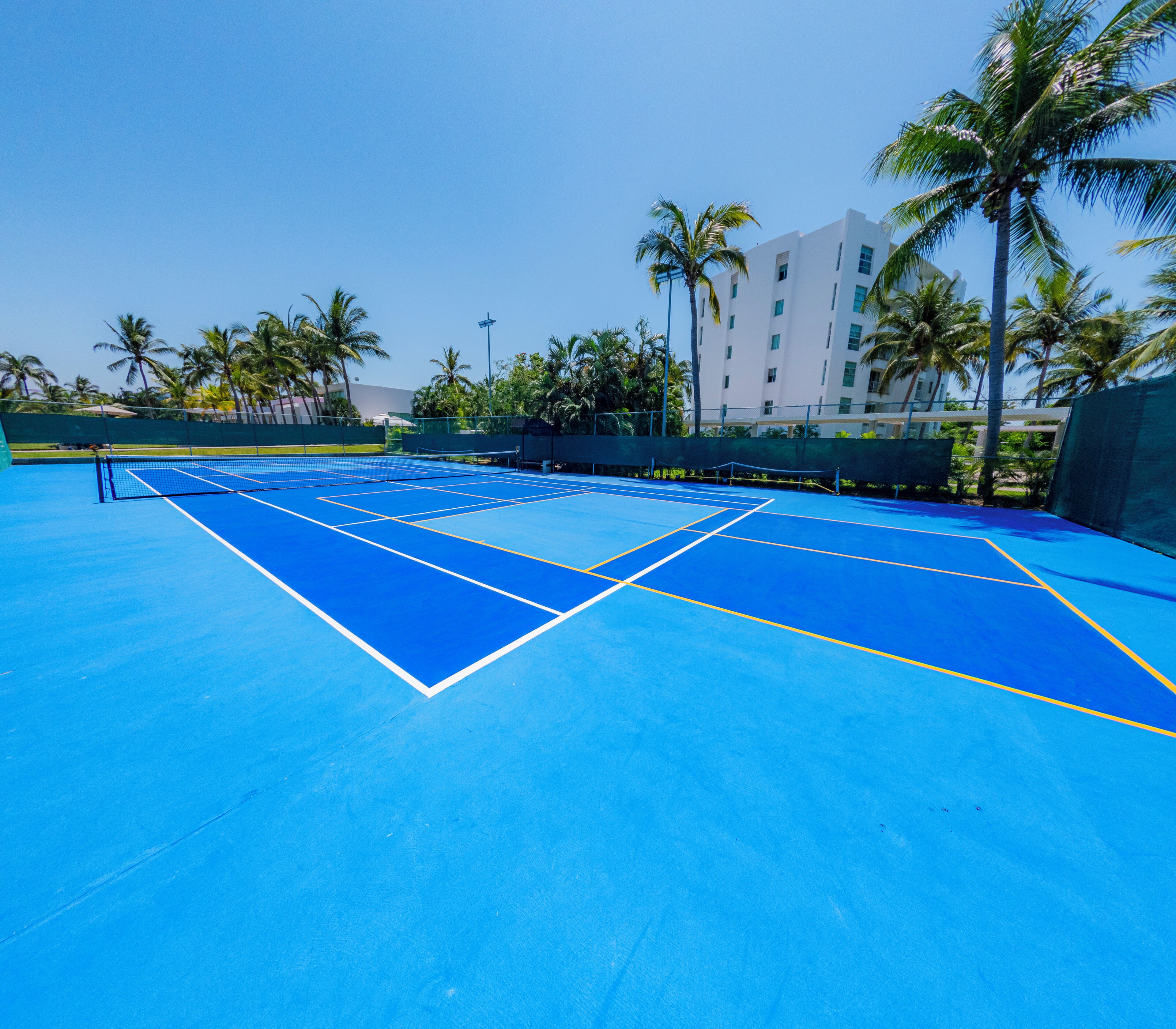 A blue tennis court with a net, surrounded by palm trees and a building in the background.