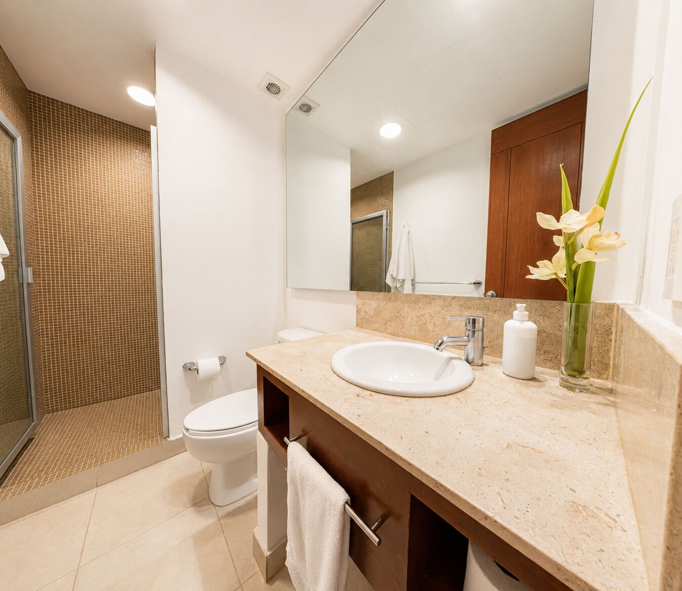 Une salle de bain moderne avec une douche en verre, des toilettes, un lavabo avec un miroir au-dessus et un vase rempli de fleurs.