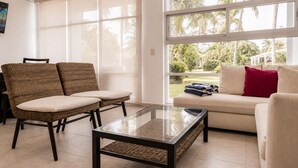 A living room with a sofa, two chairs, a glass coffee table, and large windows with a view of palm trees.