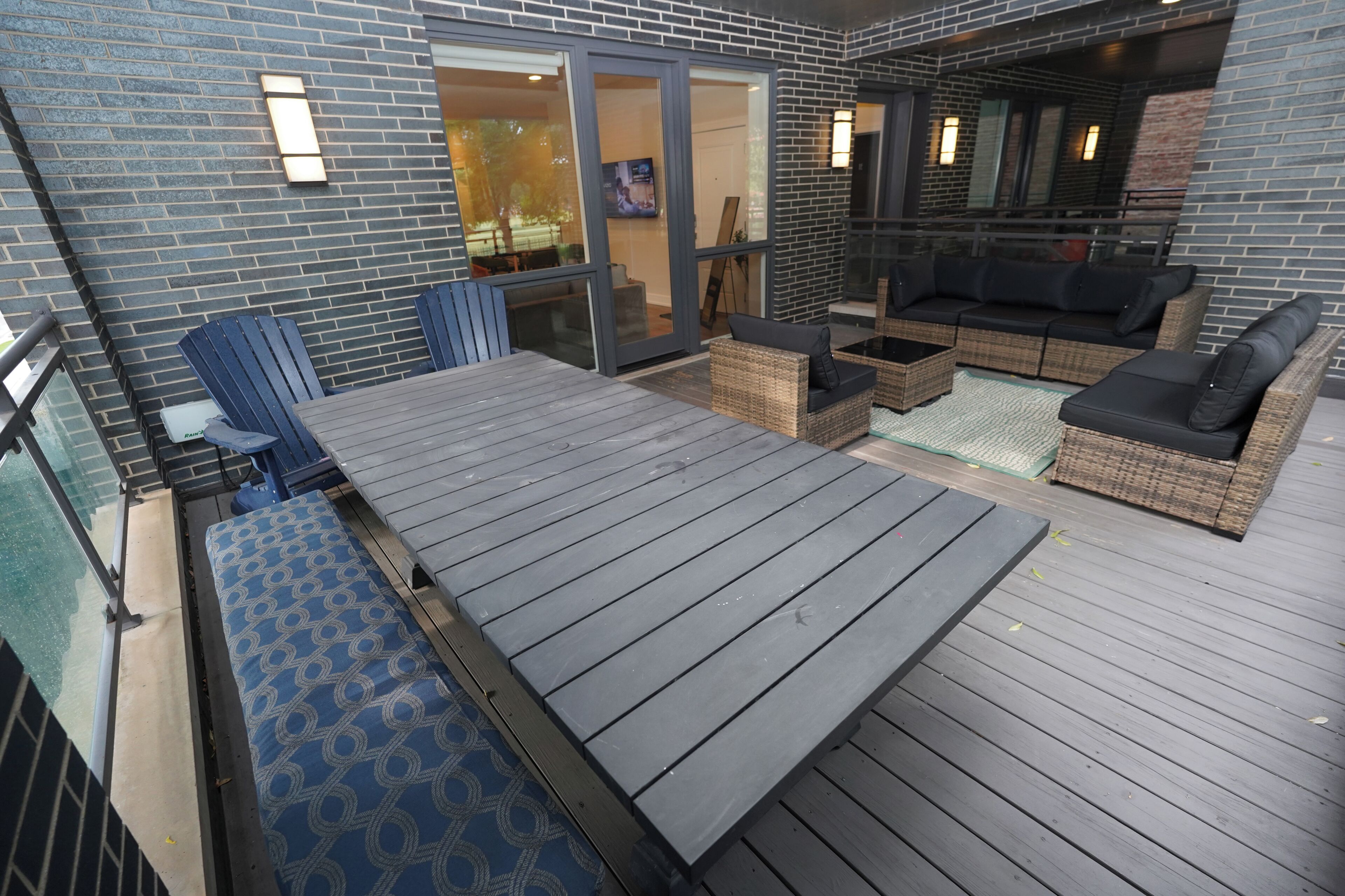 Een overdekt terras buiten met een houten vloer, rieten meubels en een glazen deur die naar een binnenruimte leidt.