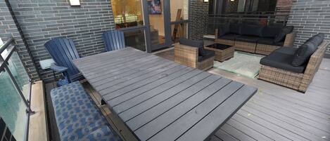 Een overdekt terras buiten met een houten vloer, rieten meubels en een glazen deur die naar een binnenruimte leidt.