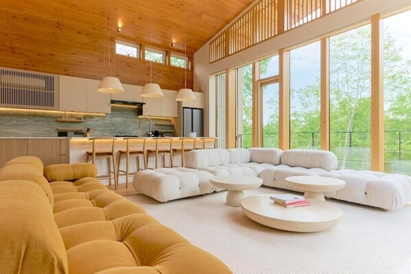 Un interior moderno con un amplio sofá seccional en forma de L, una zona de cocina y grandes ventanales que ofrecen vistas a la vegetación.
