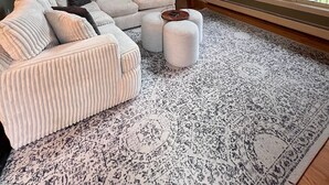 Ein geräumiges Wohnzimmer mit einer großen Eckcouch, Hocker, einem gemusterten Teppich, einer Holztreppe und einem großen Fenster mit Blick auf Bäume.