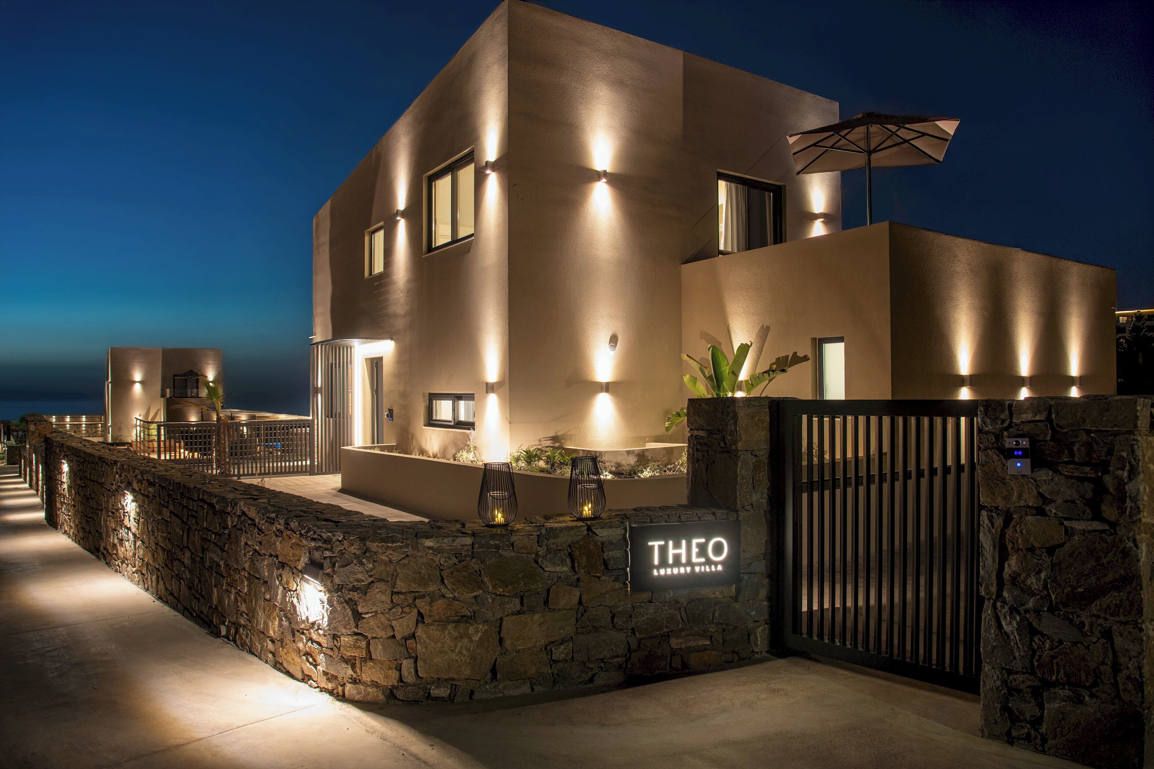 Une villa moderne avec une entrée surmontée d’un mur en pierre, une allée bordée de portails et une enseigne indiquant « VILLA DE LUXE THÉO ».