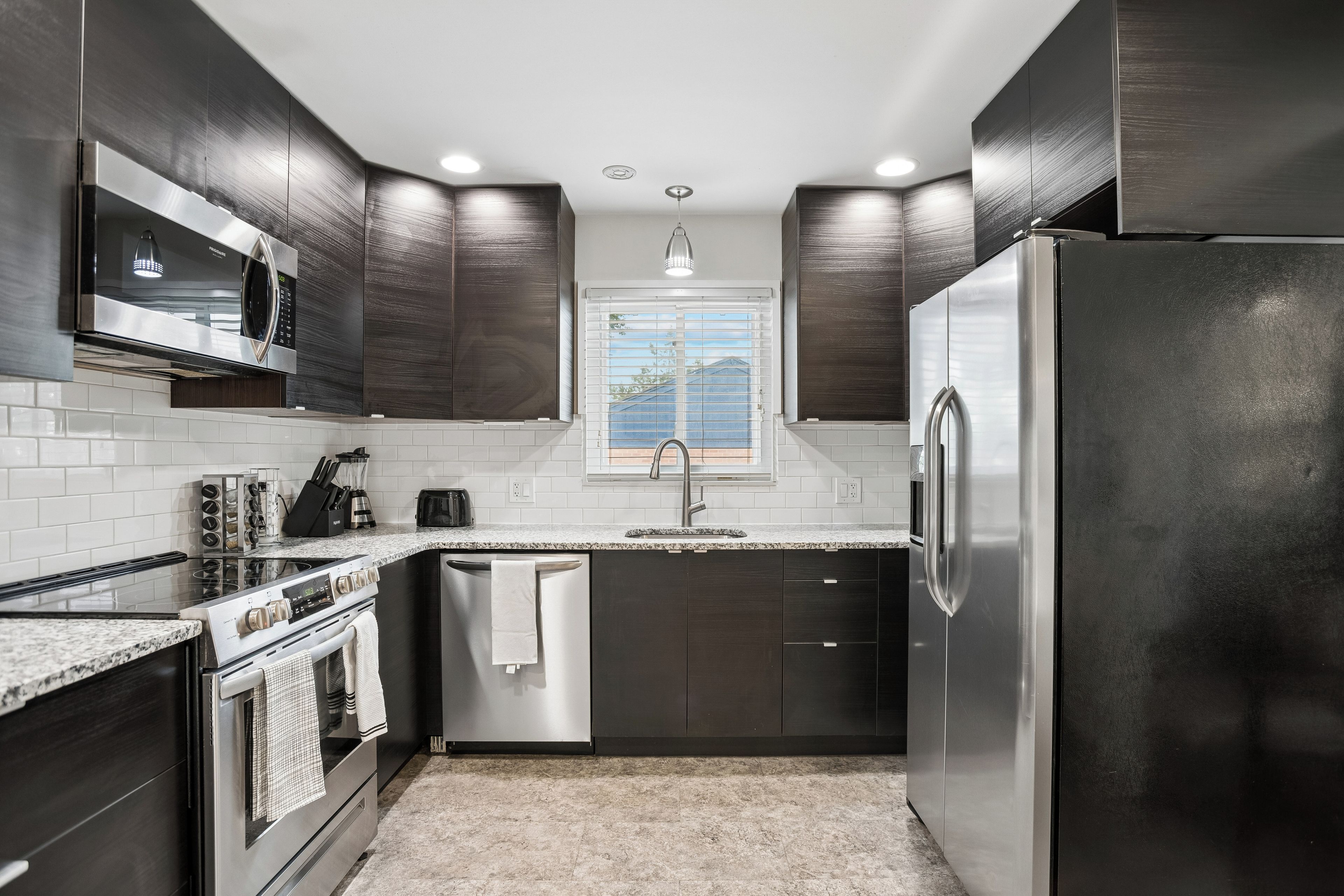 A modern kitchen with dark cabinetry, stainless steel appliances, and a window with blinds.