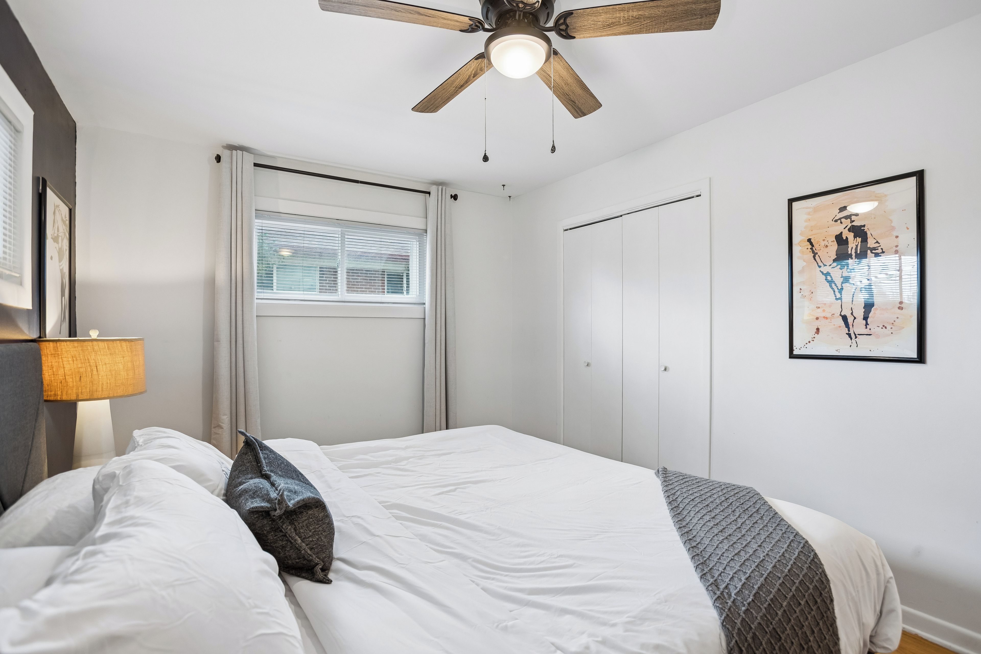 A bedroom with a bed, a ceiling fan, a window with blinds, a door, and a framed picture on the wall.