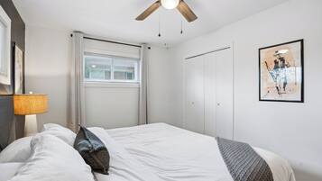 A bedroom with a bed, a ceiling fan, a window with blinds, a door, and a framed picture on the wall.