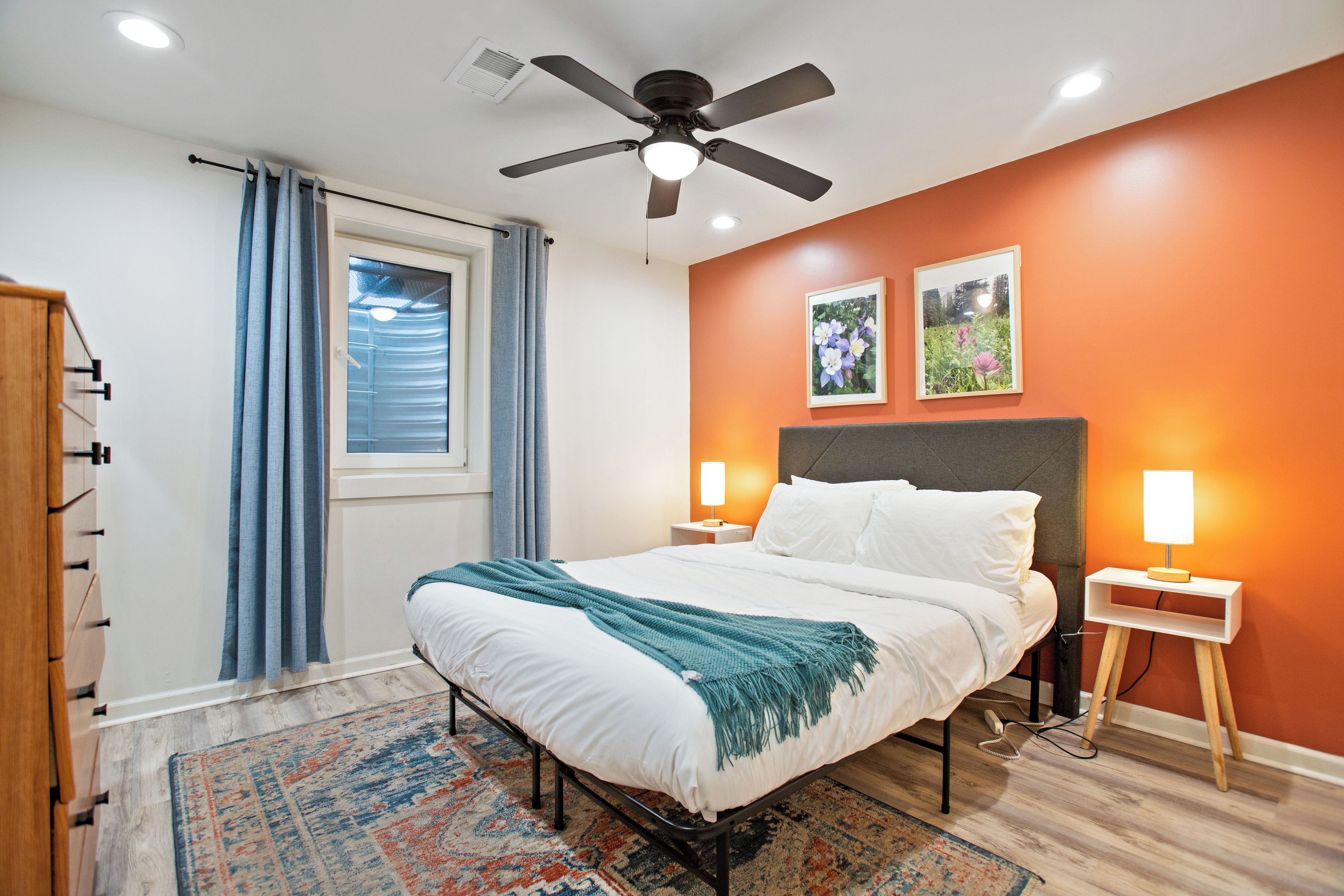 A bedroom with a bed, a ceiling fan, a window with curtains, a nightstand with a lamp, and two framed pictures on the wall.