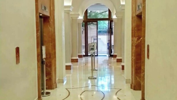 Corredor de hotel com piso de mármore, porta de entrada de vidro e placa indicando uma saída.