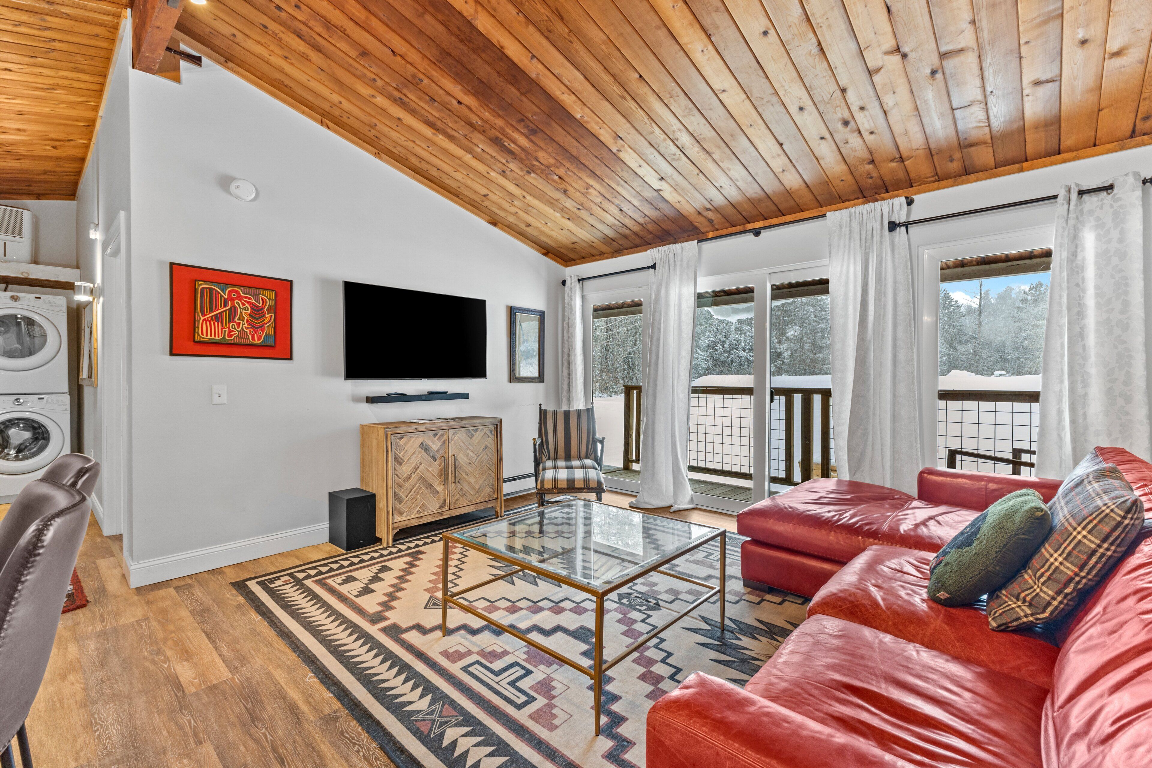 Un salon avec un canapé rouge, une table basse en verre, un plafond en bois et une grande fenêtre avec des rideaux.