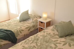 A room with two beds, each with floral-patterned bedding and pillows, a bedside table with a lit candle, and wicker baskets.