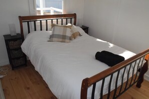 A bedroom with a wooden bed frame, white bedding, a nightstand with a lamp, and a window with a view of the outside.