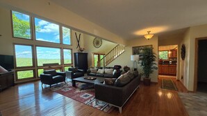 Ein geräumiges Wohnzimmer mit einer großen Couch, einem Couchtisch und einem Fernseher. Große Fenster bieten einen Blick ins Freie. Eine Treppe führt zu einer weiteren Ebene.
