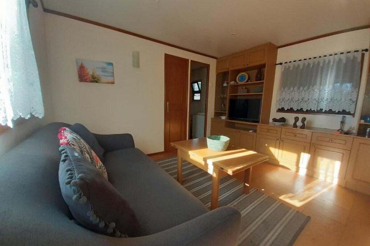 Sala de estar com sofá, uma mesa pequena e área de cozinha com armários e janela.