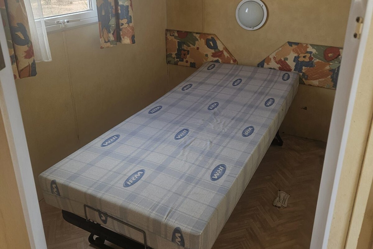 Quarto pequeno com cama de solteiro, janela, luminária na parede e cortina estampada.