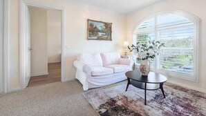 Una habitación con un sofá blanco, una mesa redonda con un florero, un cuadro en la pared y un ventanal grande.