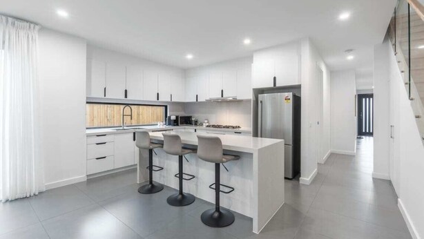 Una cocina moderna con muebles blancos, una isla central con taburetes altos, un refrigerador y un microondas.