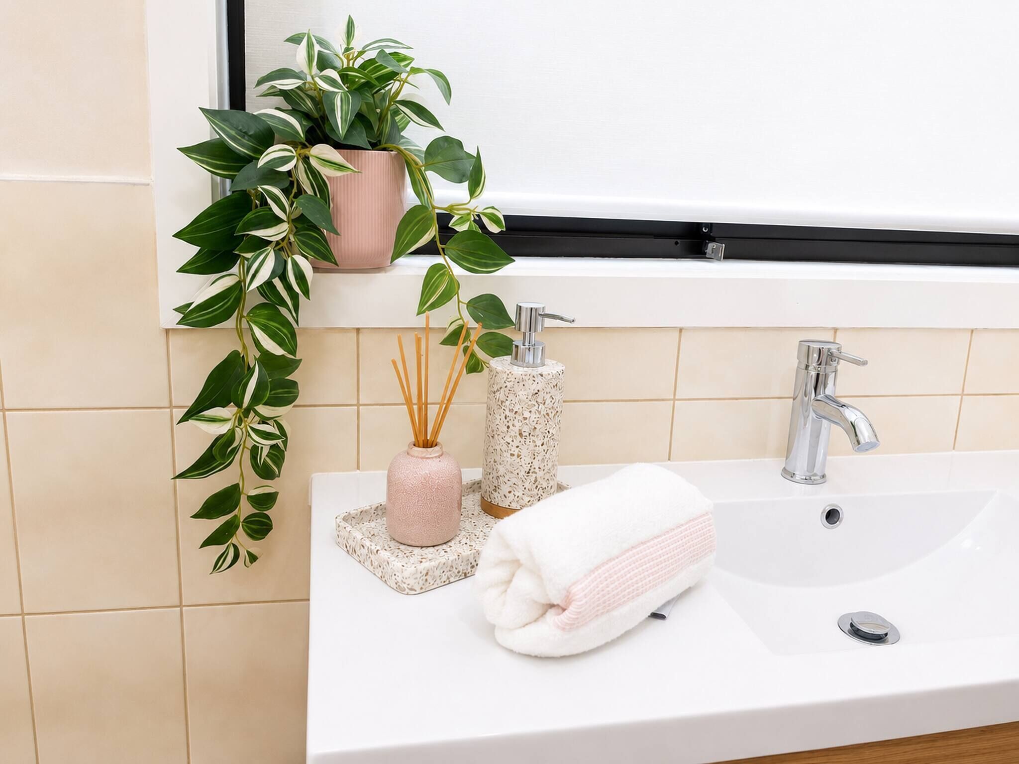 A bathroom with a white sink, two faucets, a towel, a pink vase with reed sticks, and a green plant.