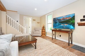 Ein Wohnzimmer mit einem Glastisch, einer Couch, einem Fernseher auf einem Holzboden und einer Treppe sowie einer Tür.