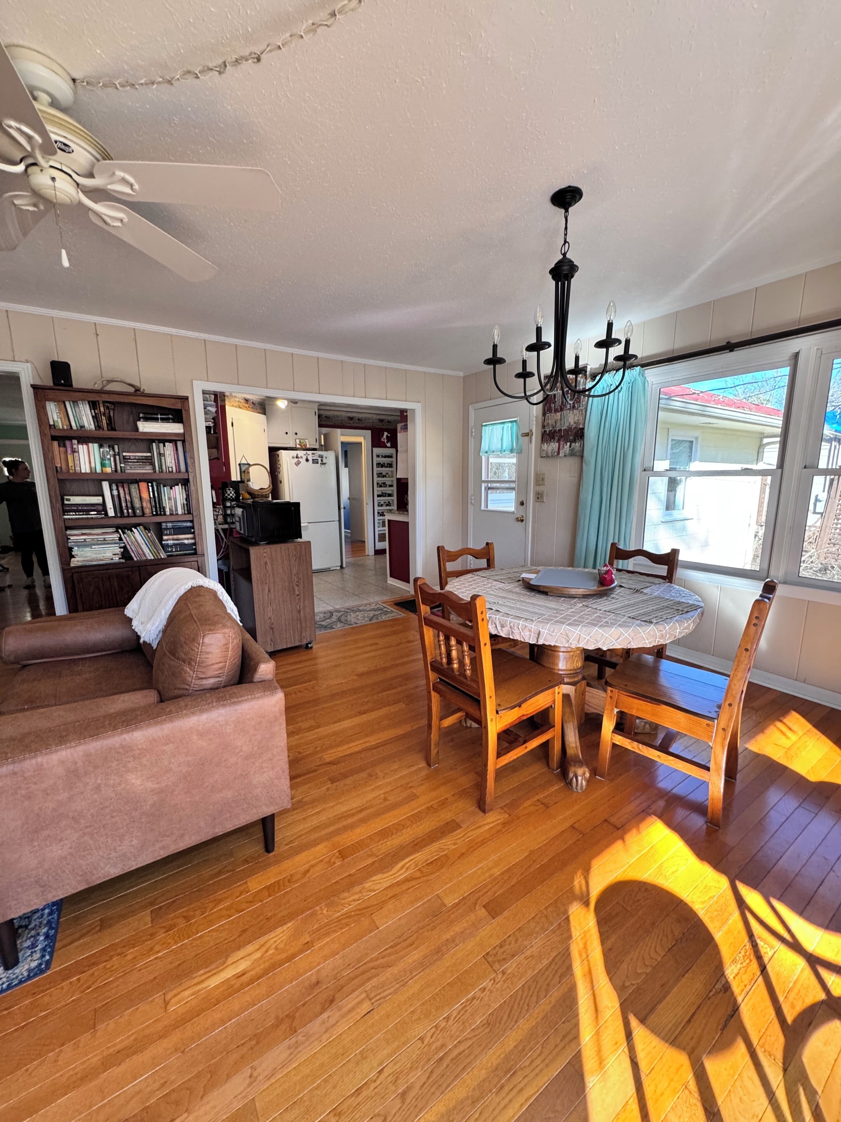 A spacious living room with a wooden floor, a dining table with chairs, a sofa, and a ceiling fan.
