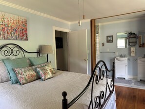 A bedroom with a bed, a nightstand, a lamp, a painting, a bathroom with a sink and mirror.
