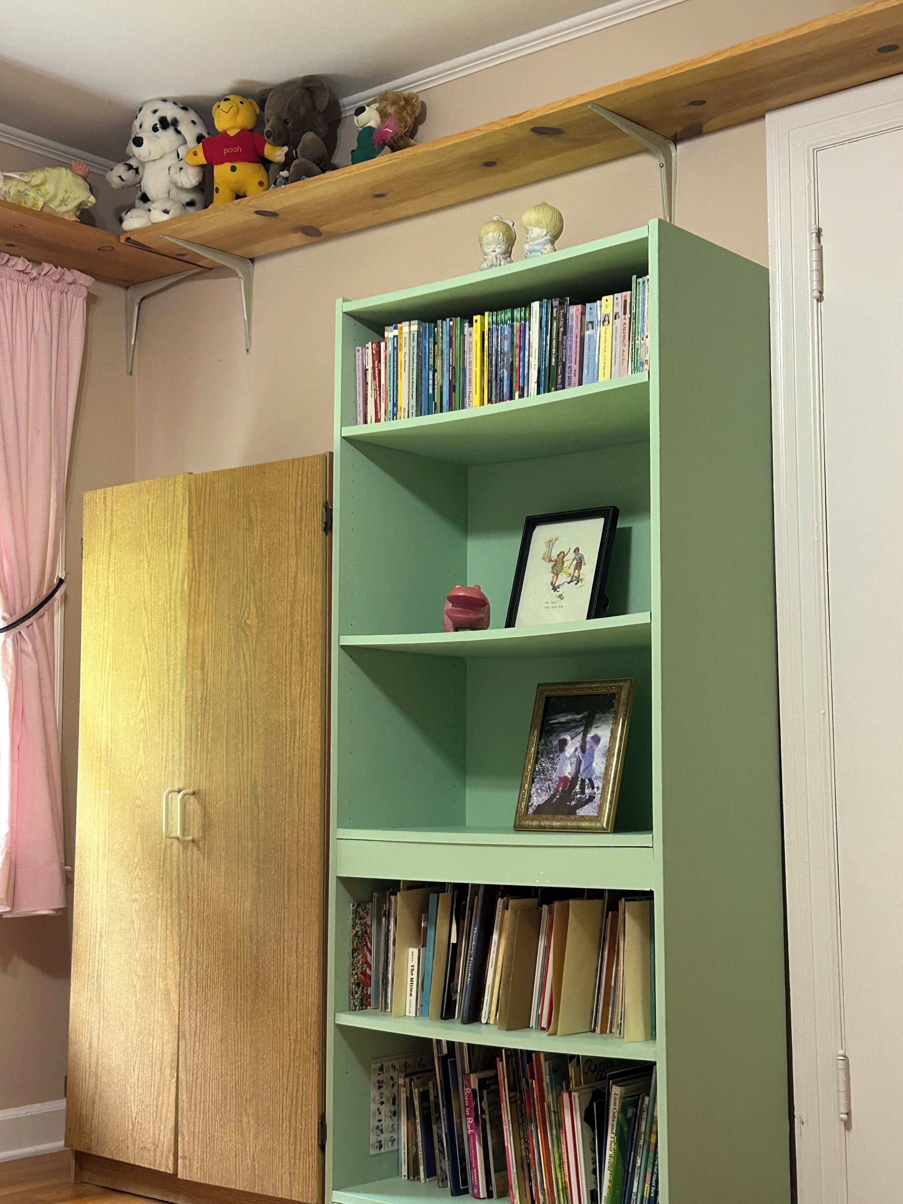 A room with a green bookshelf filled with books, a wooden wardrobe, and a pink curtain.