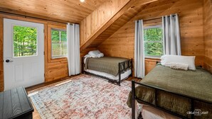 Eine Holzhütte mit zwei Betten, einer Holzbalkendecke und Blick auf den Wald durch die Fenster.