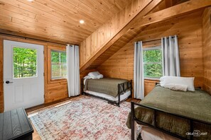 Eine Holzhütte mit zwei Betten, einer Holzbalkendecke und Blick auf den Wald durch die Fenster.