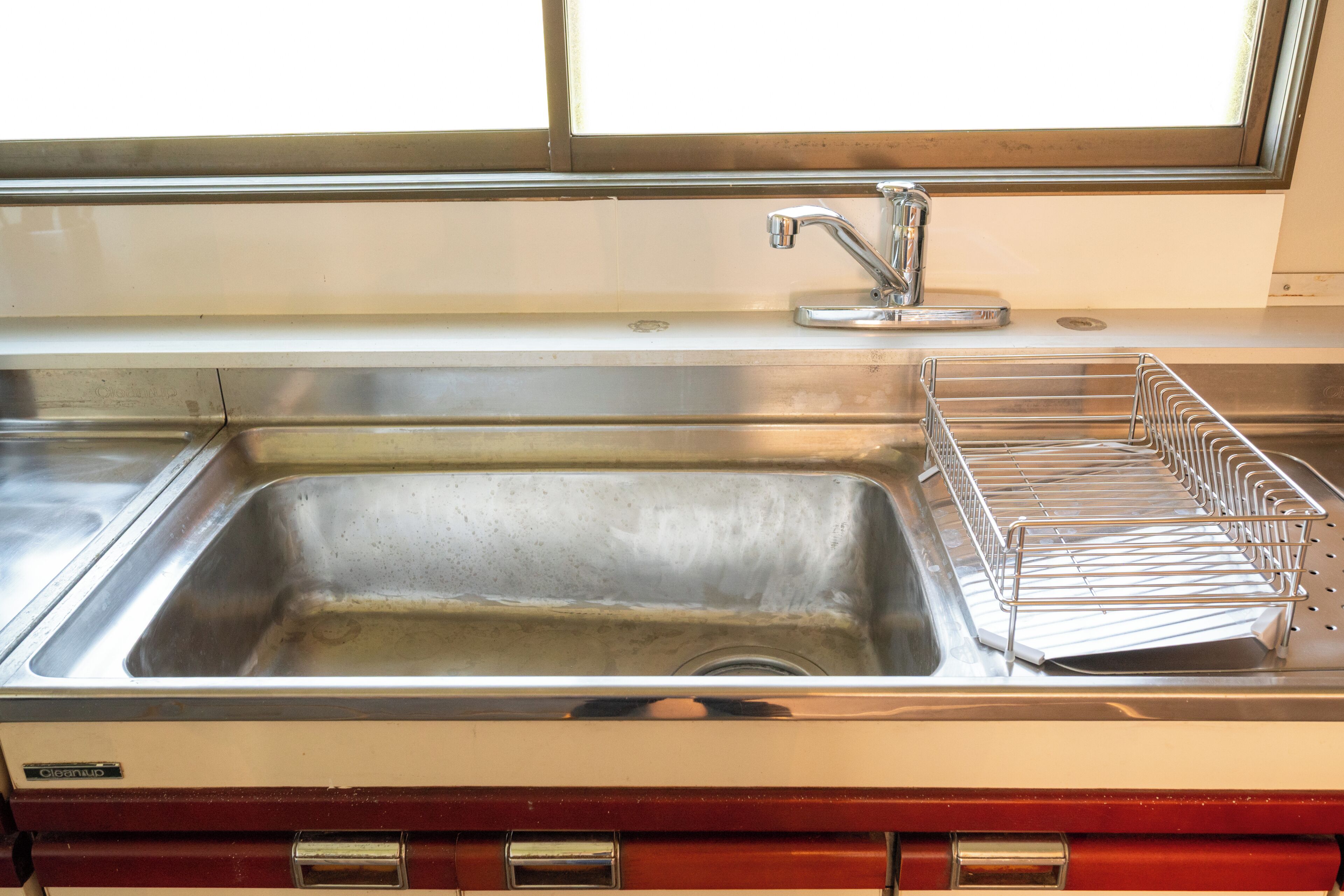 A stainless steel sink with a chrome faucet, a metal drying rack, and a window above the sink.