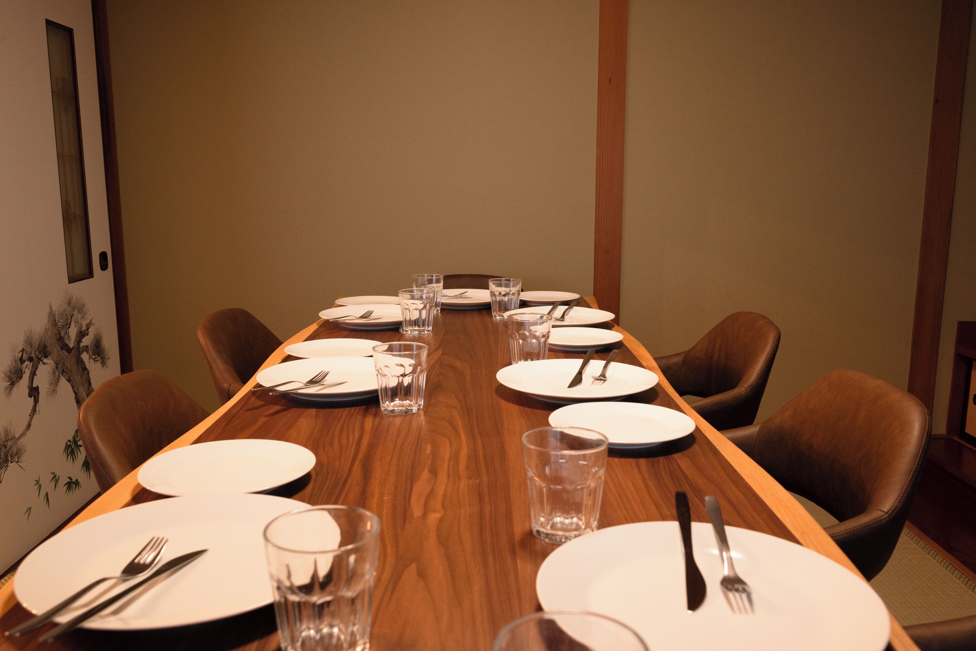 Una larga mesa de madera dispuesta para una comida, con platos blancos, cubiertos y vasos de vidrio.