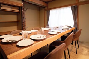 A dining area with a long wooden table set for a meal, chairs, and a window with curtains. - Kamifurano Cozy House (Kamifurano-cho, Sorachi District)