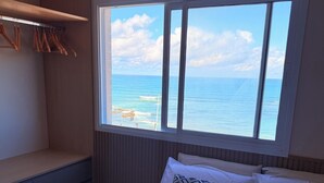 A bedroom with a bed, pillows, a window with a view of the sea, a bench, and a wooden wall. - Beachfront Studio in Piatã | Incredible View, Wi-Fi, Smart TV and Garage (Salvador)