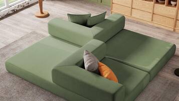 Un sofá seccional verde con cojines, una mesita auxiliar con una planta y un estante de madera con libros y objetos decorativos.