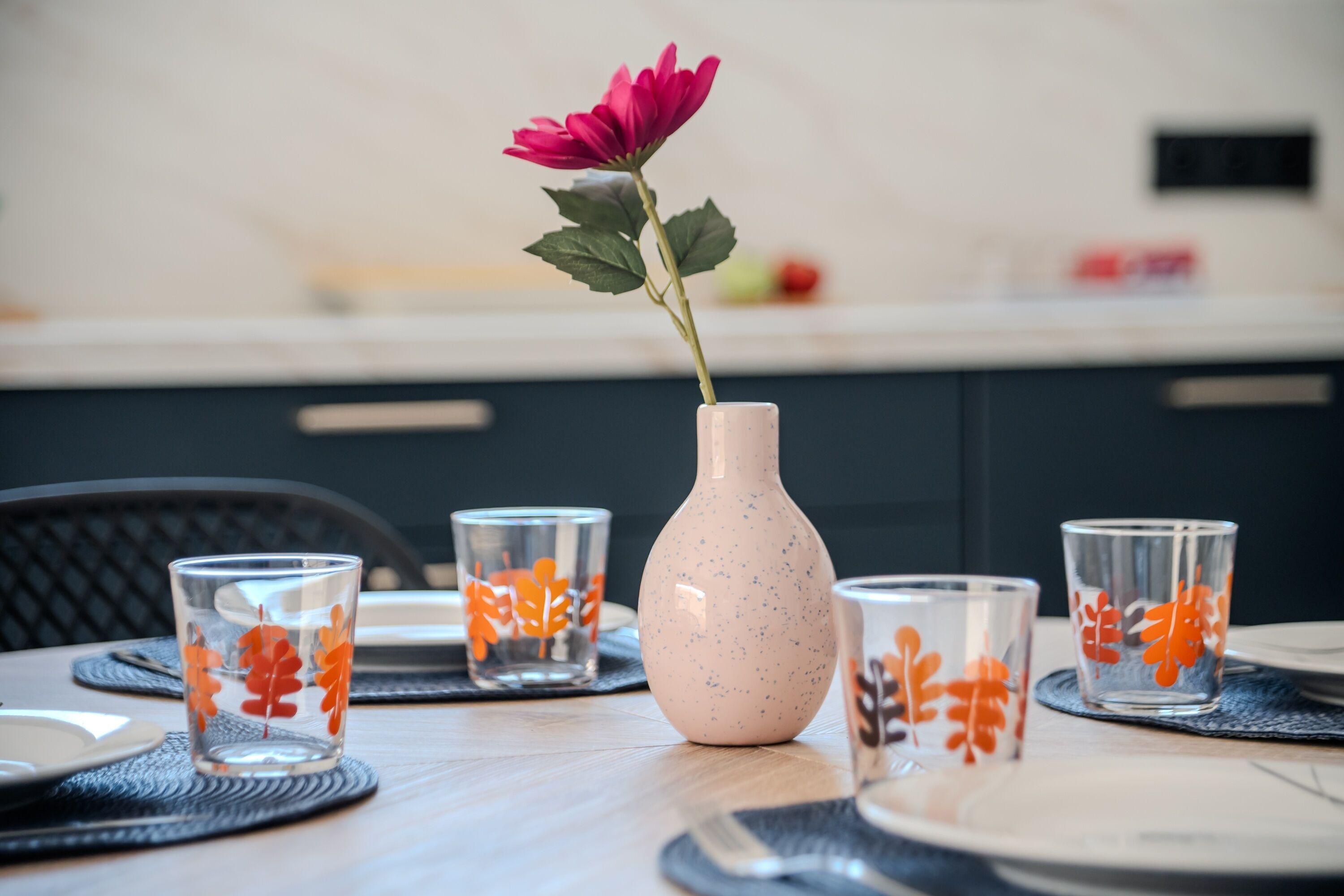 A dining table with a vase of flowers, clear glasses with leaf patterns, and a modern kitchen in the background.