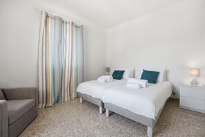 A double bed with white bedding, a grey bench, a striped curtain, and a white nightstand with a lamp. - Le Mas des Aromes - Piscine Privée (Mandelieu-la-Napoule)