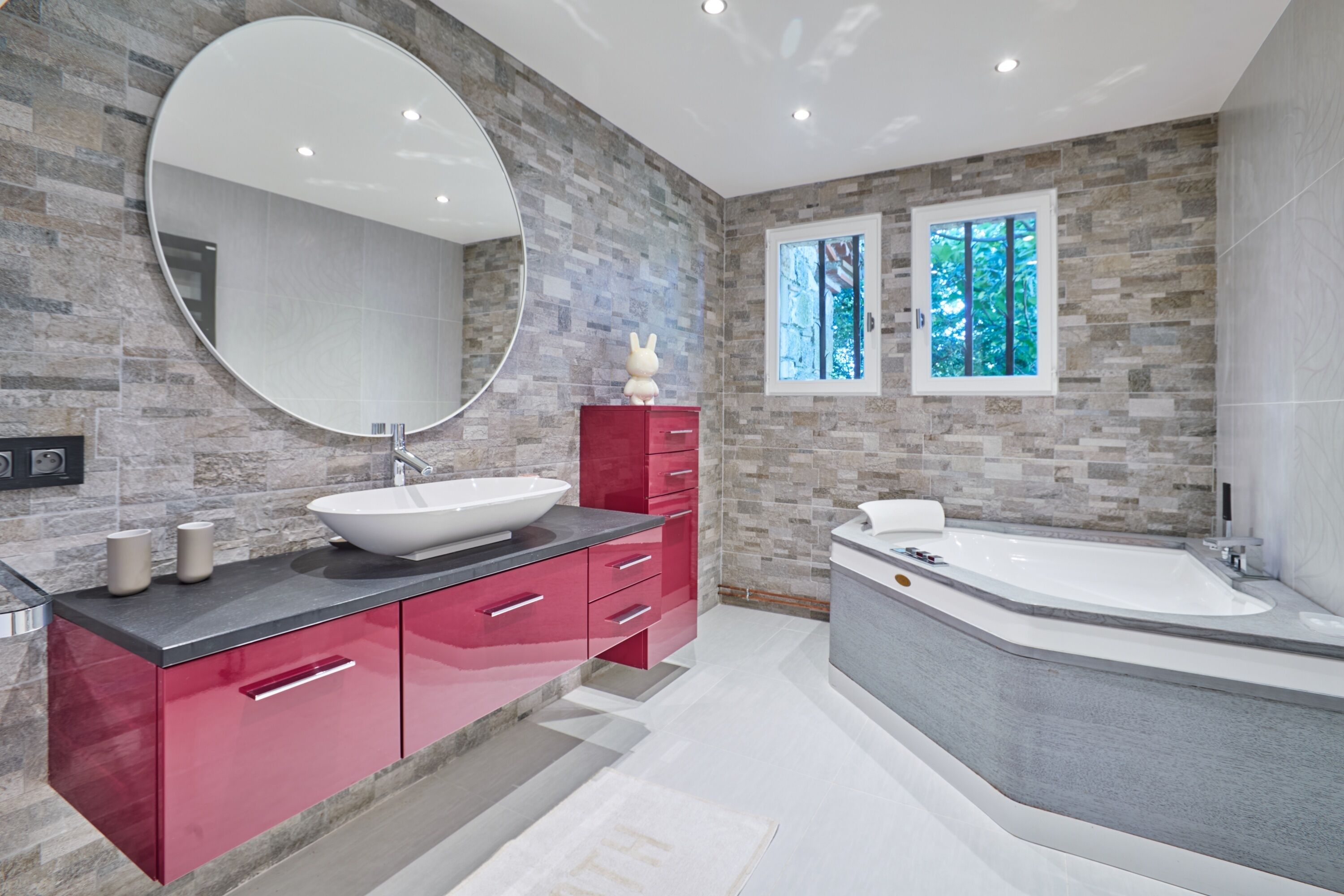 A modern bathroom with a large round mirror, a vessel sink, a red vanity, and a freestanding bathtub.