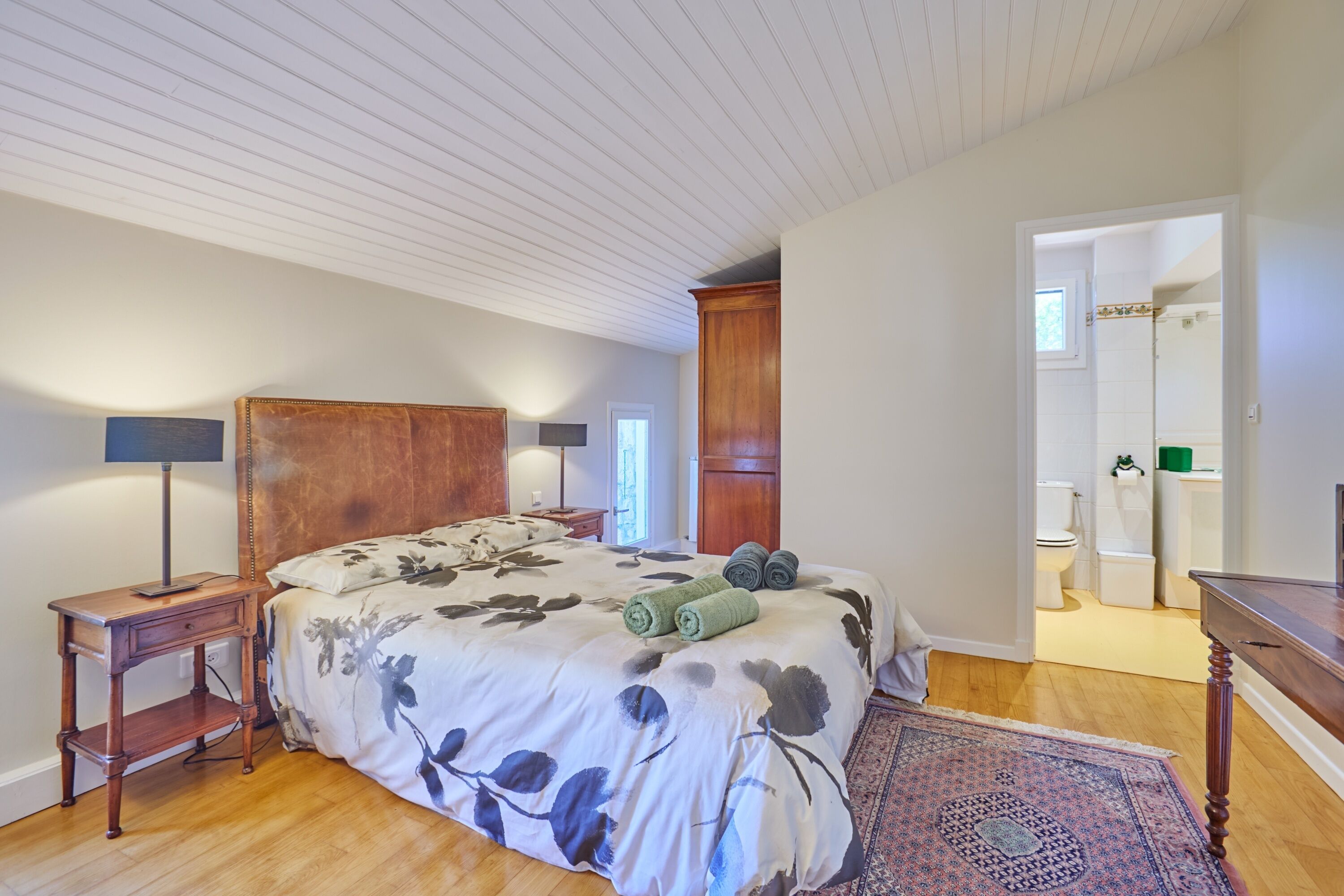 A bedroom with a large bed, wooden headboard, bedside lamps, a patterned rug, and a view of the bathroom.